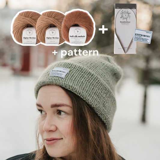 Woman wearing a green knit beanie with yarn packages and a pattern book in the background.