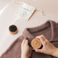 Person holding a wooden tag on a brown garment with a small brush and white bag in the background.