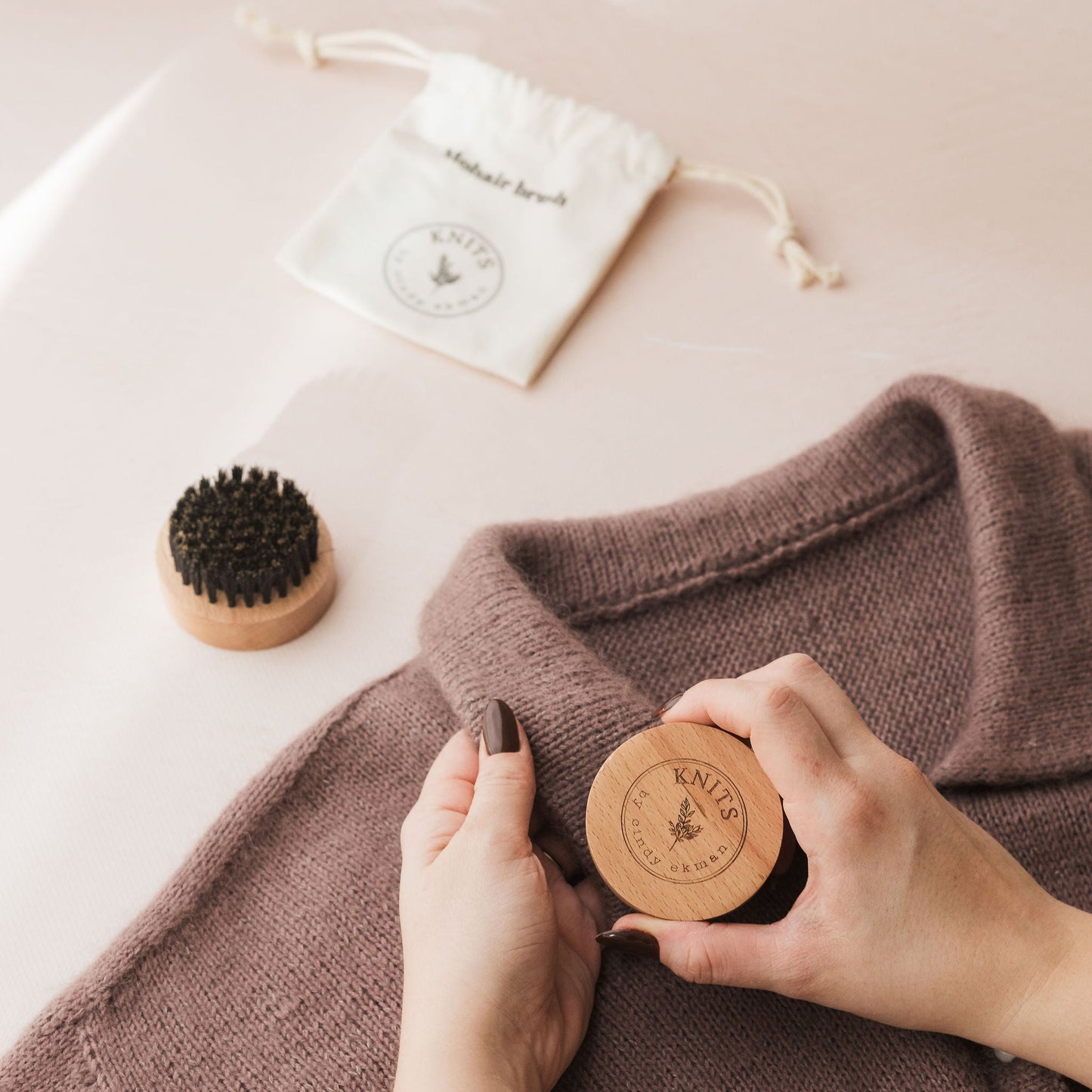 Person holding a wooden tag on a brown garment with a small brush and white bag in the background.