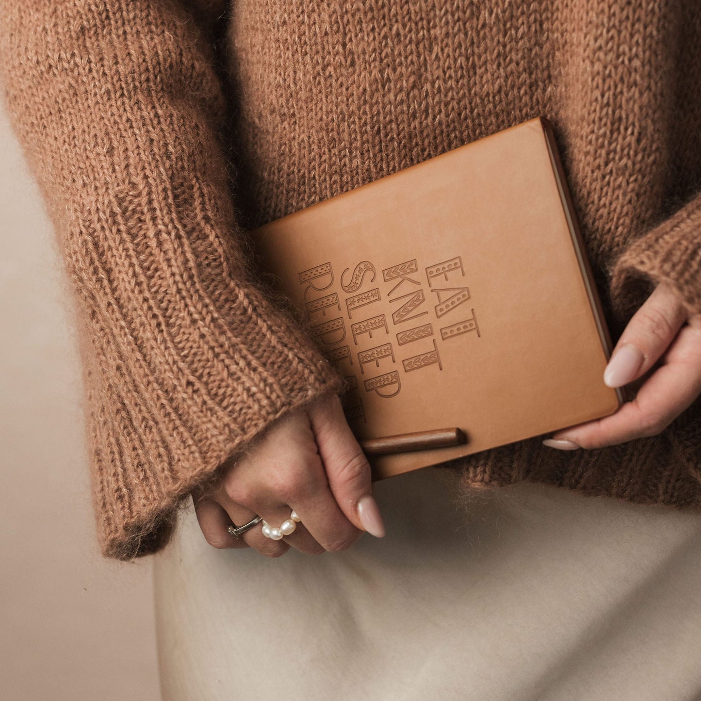 Person wearing a brown sweater holding a brown leather notebook with 'EAT KNIT SLEEP' text.