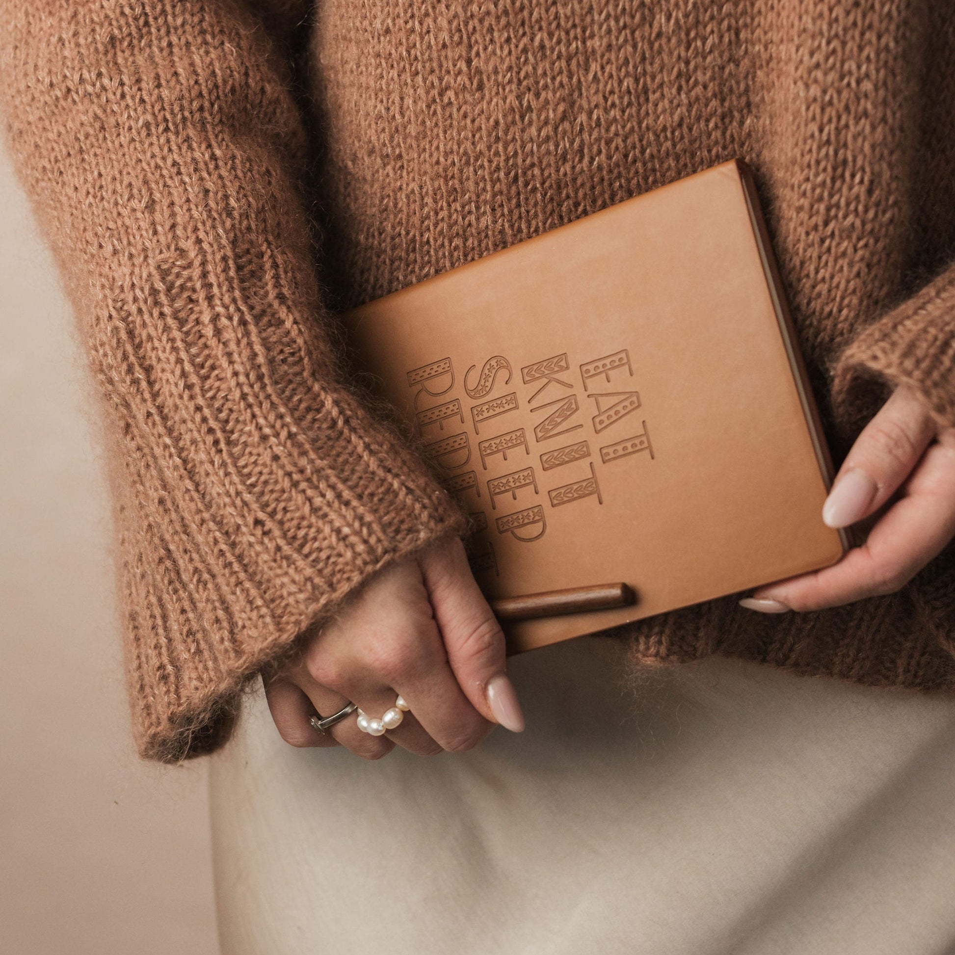 Person wearing a brown sweater holding a brown leather notebook with 'EAT KNIT SLEEP' text.