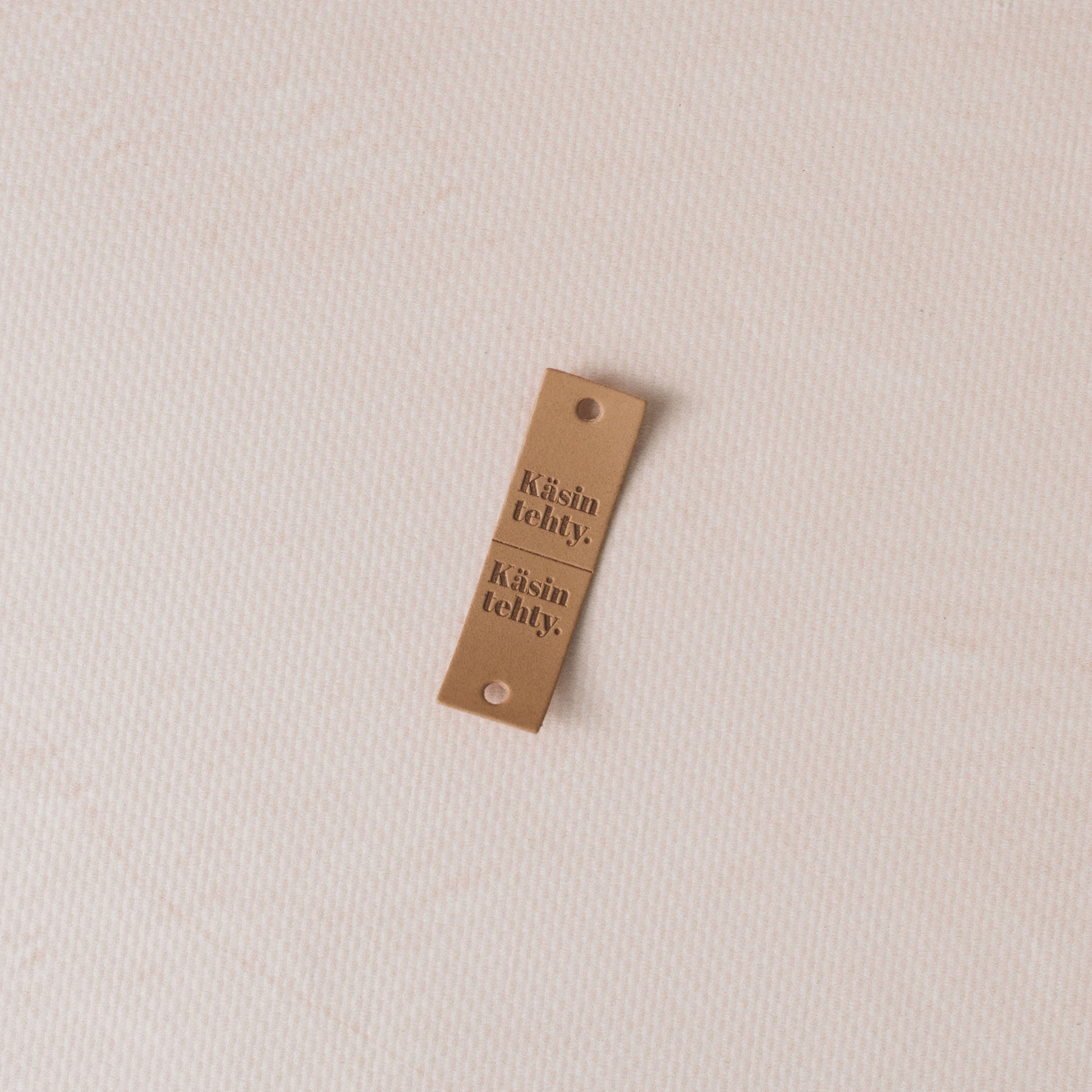 PU leather paper tag with text "käsin tehty" on a light beige fabric background