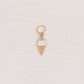 A small ice cream cone charm with white enamel and a tiny green gem, attached to a gold ring, displayed on a textured off-white background.
