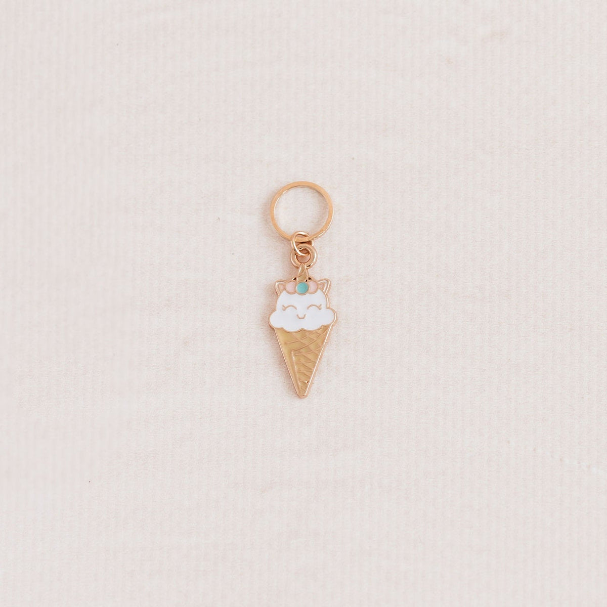 A small ice cream cone charm with white enamel and a tiny green gem, attached to a gold ring, displayed on a textured off-white background.