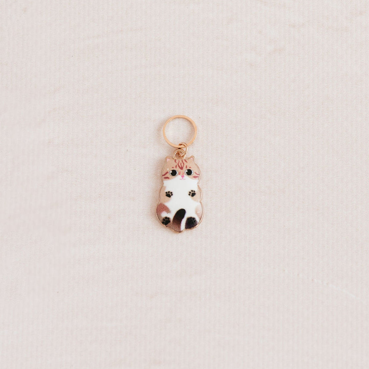 A small rose gold charm shaped like a cute white cat wearing a pink crown, resting on a textured off-white surface.