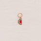 A small watermelon slice charm with a rose gold metal border and loop, featuring red, green, and white enamel details, placed on a textured off-white background.