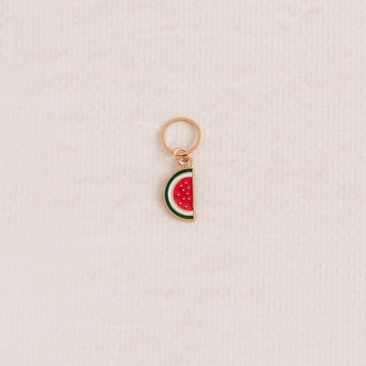 A small watermelon slice charm with a rose gold metal border and loop, featuring red, green, and white enamel details, placed on a textured off-white background.