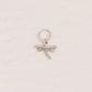 A small silver dragonfly charm with a textured body and wings, attached to a matching silver jump ring, displayed on a textured off-white background.