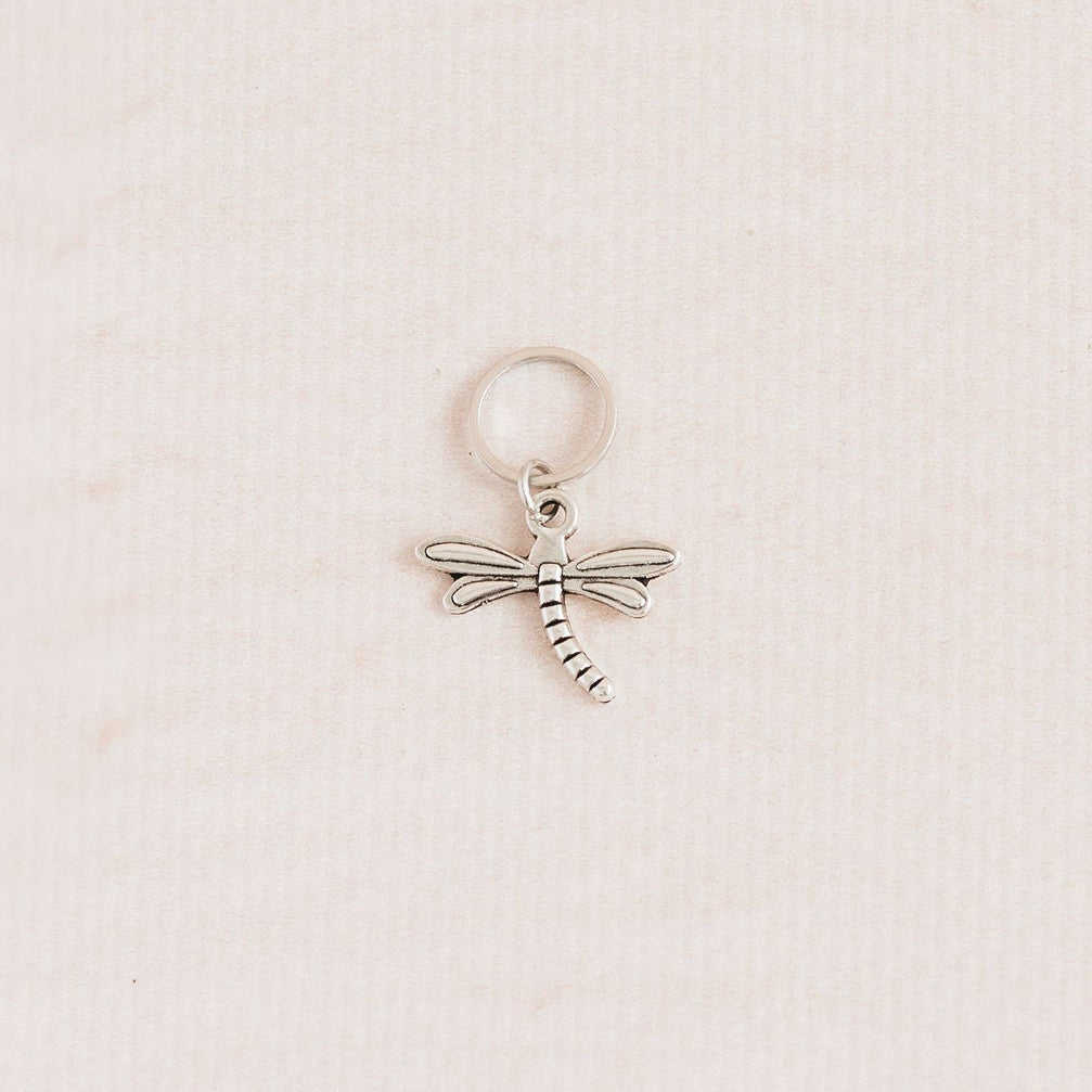 A small silver dragonfly charm with a textured body and wings, attached to a matching silver jump ring, displayed on a textured off-white background.