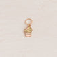 A small rose gold cupcake charm with a green frosting swirl and pink heart accent, displayed on a textured cream background.