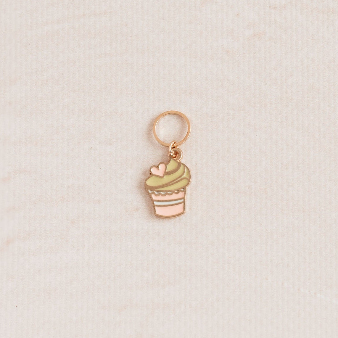 A small rose gold cupcake charm with a green frosting swirl and pink heart accent, displayed on a textured cream background.