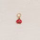 A small ladybug charm with red and white enamel wings and gold-tone metal trim, hanging from a gold ring on a textured off-white background.