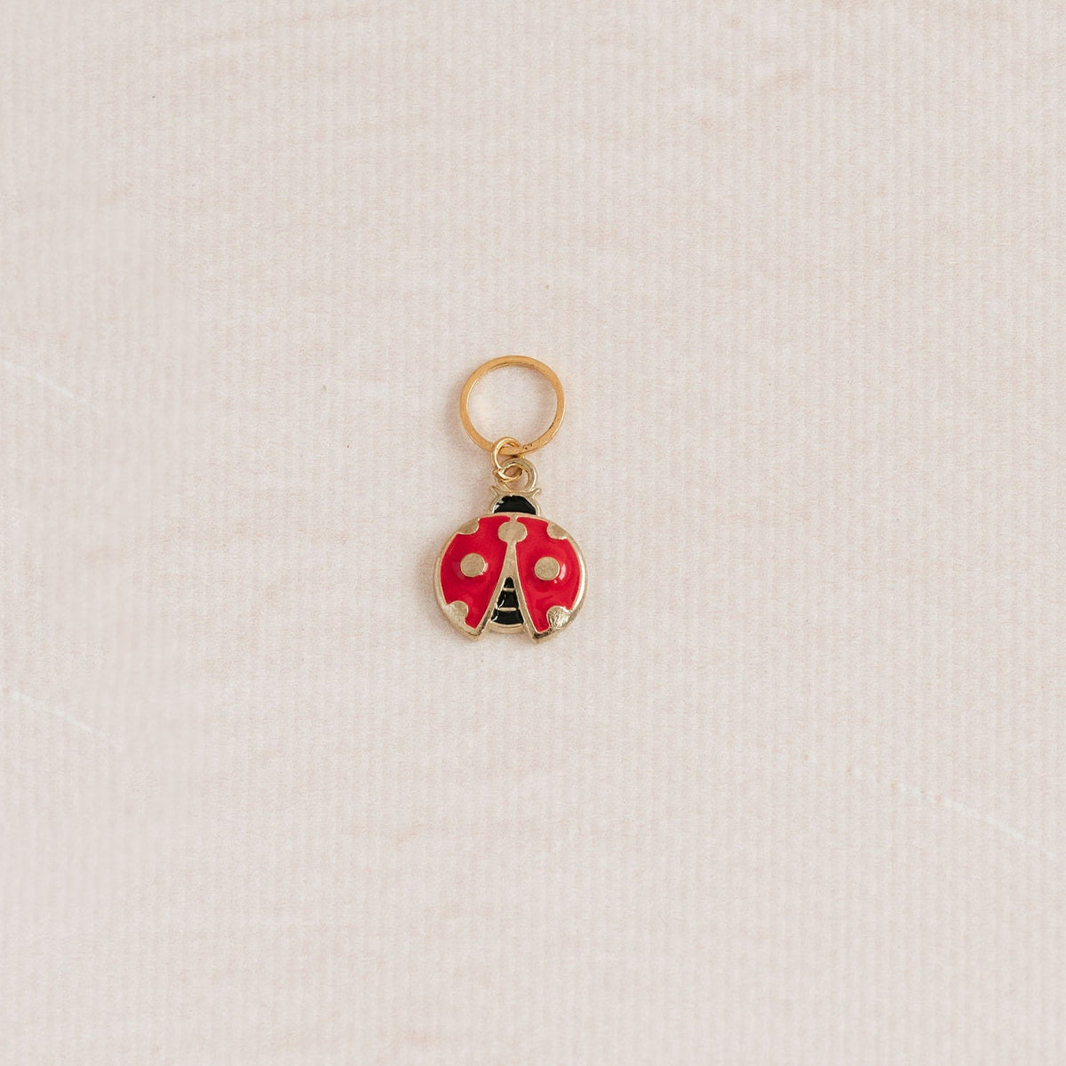 A small ladybug charm with red and white enamel wings and gold-tone metal trim, hanging from a gold ring on a textured off-white background.