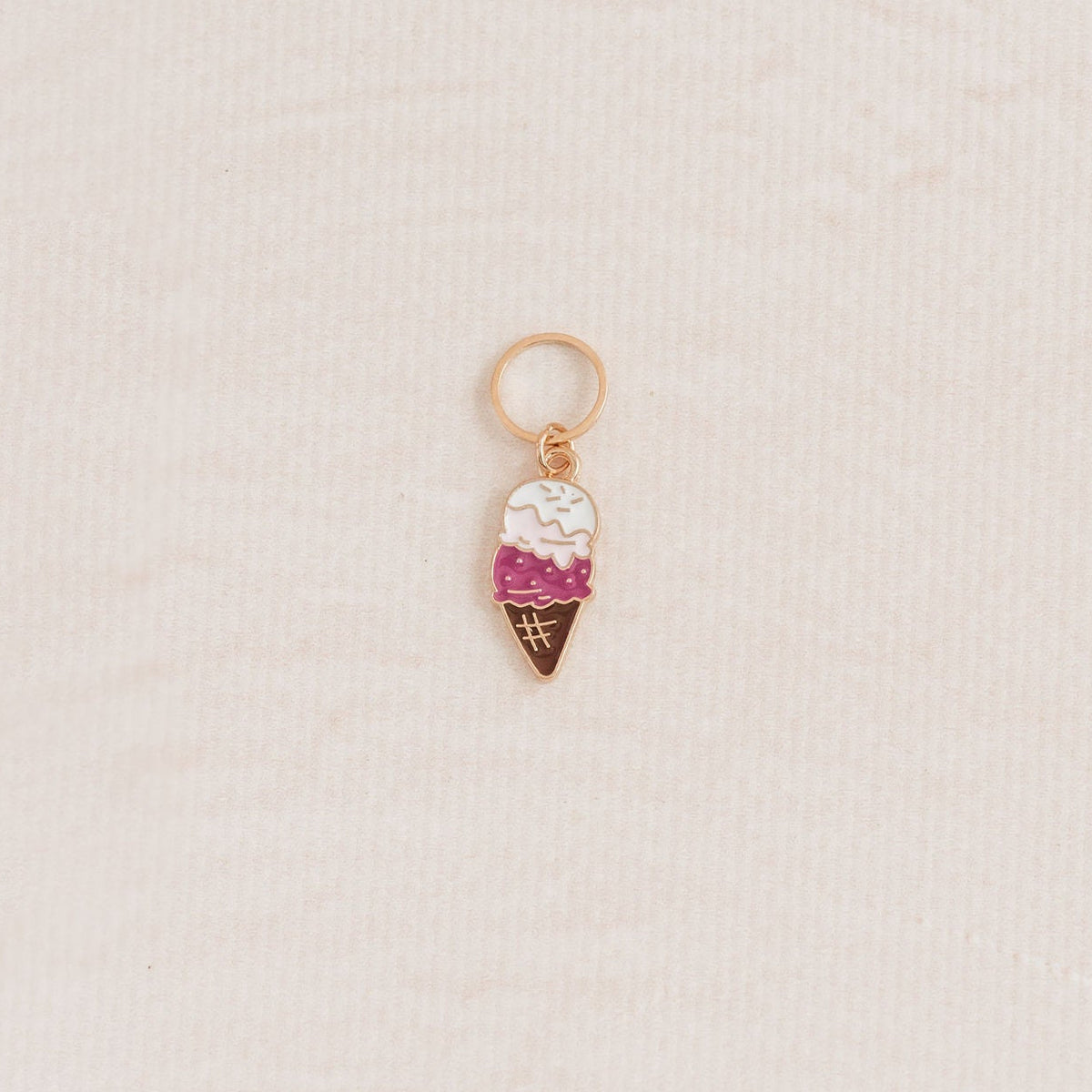 A small ice cream cone charm with a gold-tone ring at the top, featuring white and pink enamel detailing, placed on a textured off-white background.