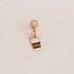 A small gold-tone charm shaped like a takeaway coffee cup with white and brown enamel details and the word 'coffee' printed on it, shown on a light beige textured background.