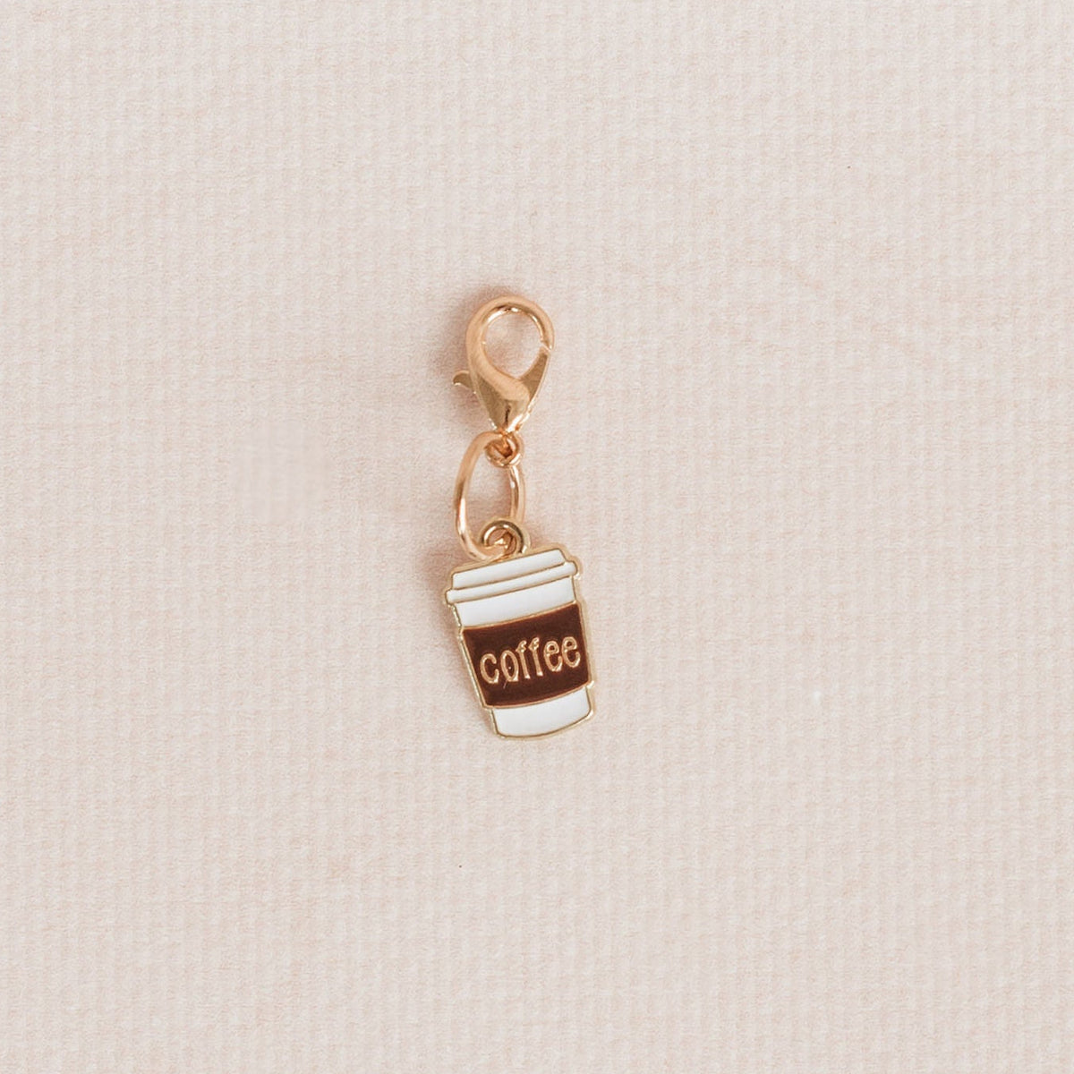 A small gold-tone charm shaped like a takeaway coffee cup with white and brown enamel details and the word 'coffee' printed on it, shown on a light beige textured background.