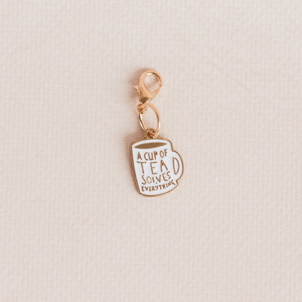 A small gold charm shaped like a teacup with the text 'A CUP OF TEA SOLVES EVERYTHING" in gold lettering, shown on a textured off-white background.