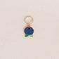 A small gold-tone charm shaped like a blueberry with a dark blue enamel body, a tiny white star detail, and green leaf accents, hanging from a gold ring on a textured off-white background.