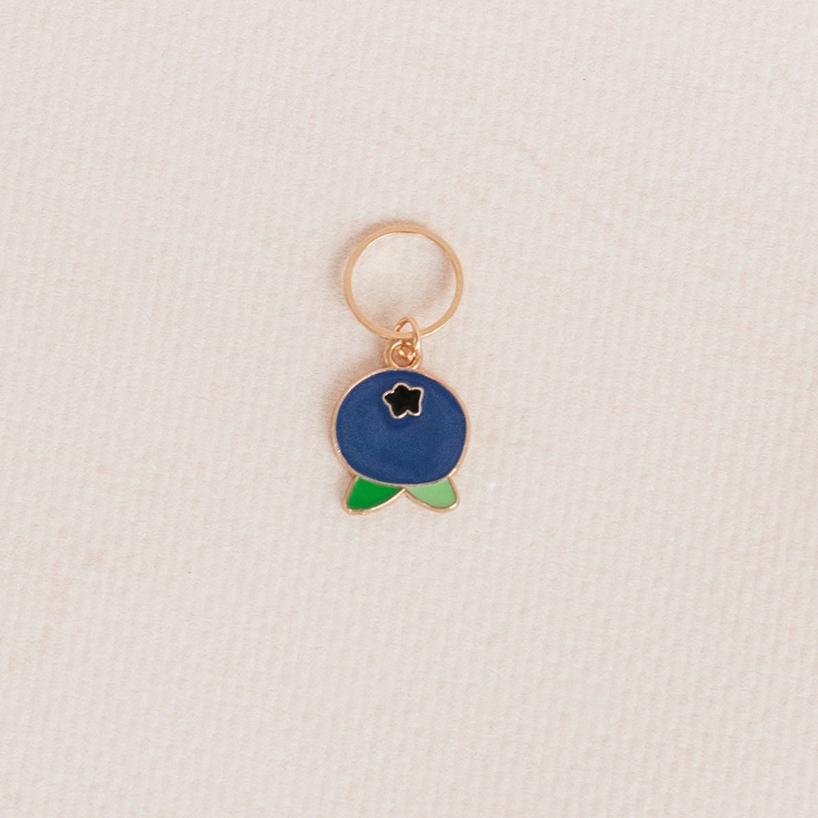 A small gold-tone charm shaped like a blueberry with a dark blue enamel body, a tiny white star detail, and green leaf accents, hanging from a gold ring on a textured off-white background.