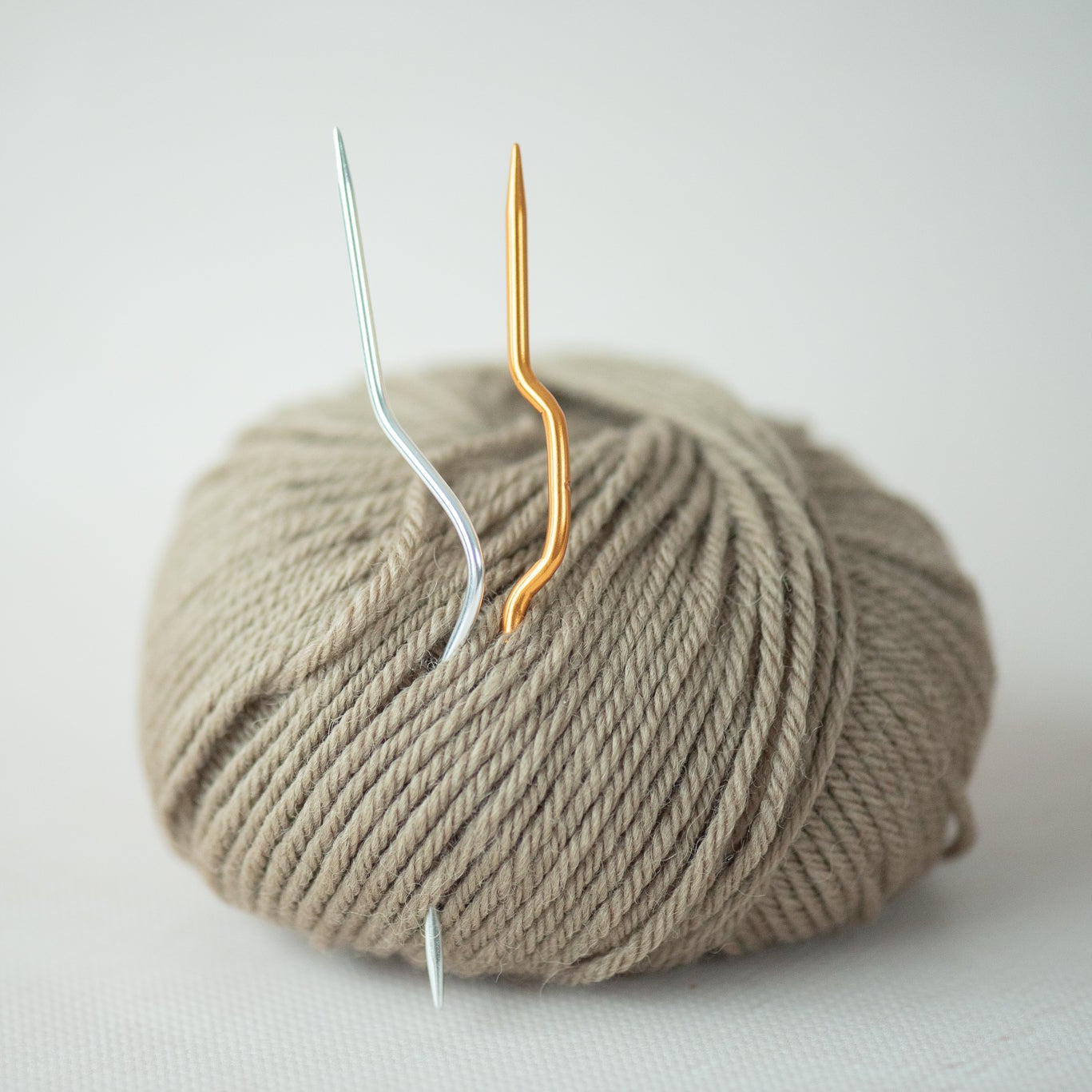 Ball of beige yarn with two metal hooks on a light gray background